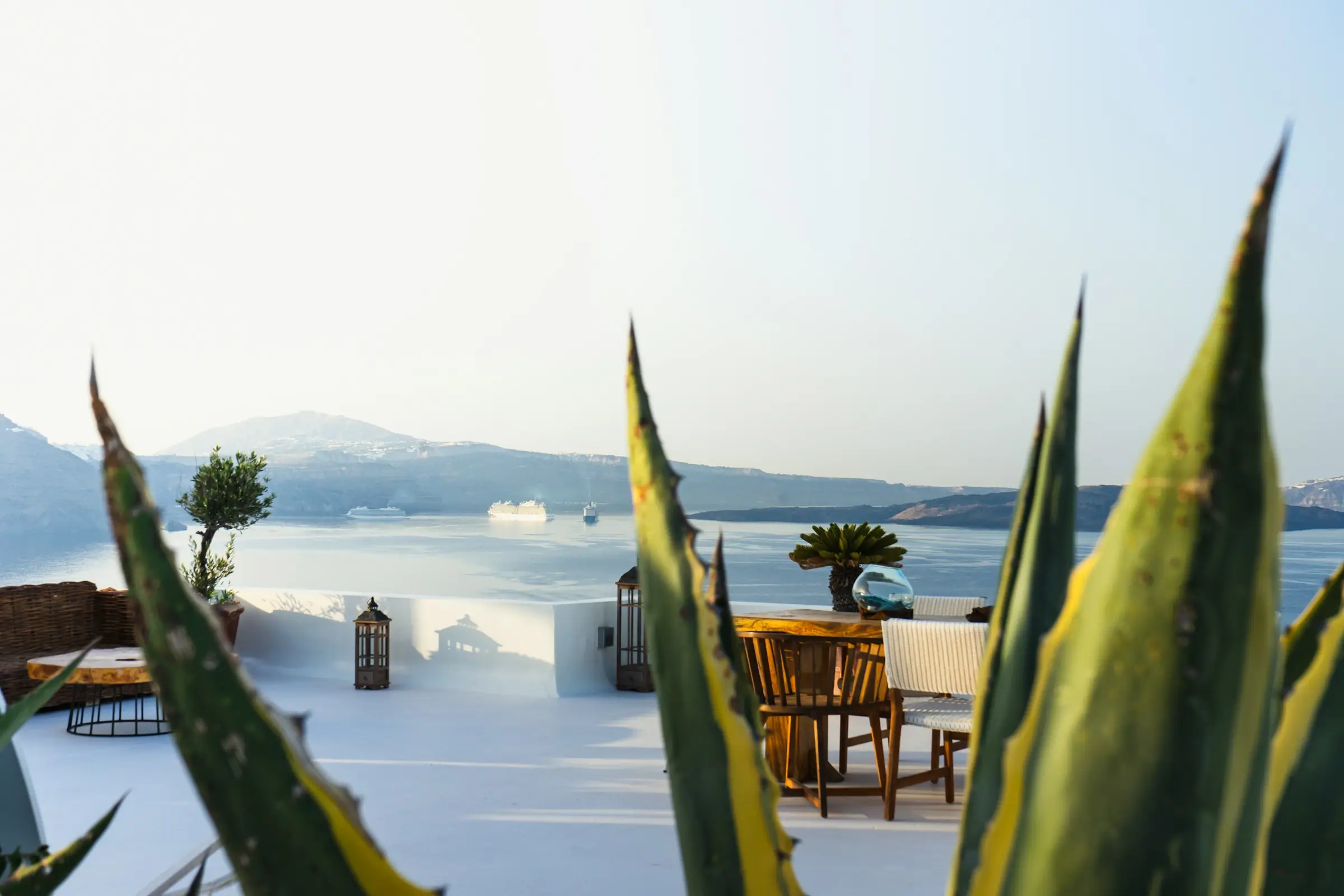 Santorini terrace with wooden table and chairs, agave leaves in front, caldera sea view
