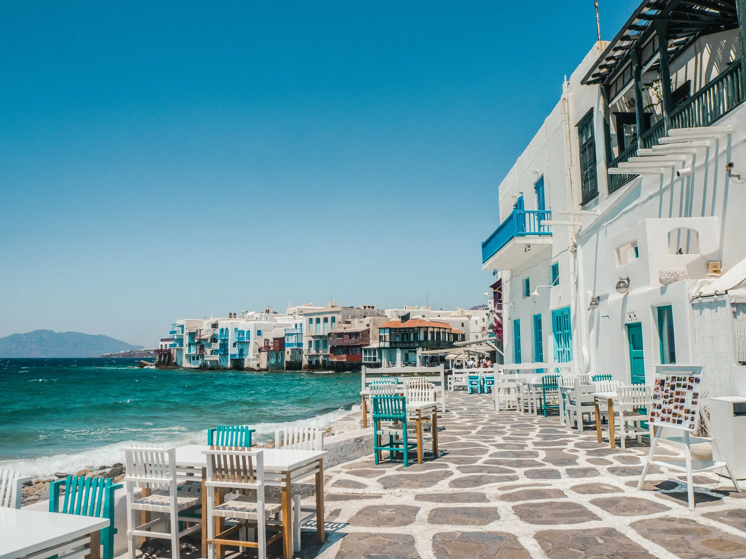 White tables and turquoise chairs on a stone waterfront terrace in Little Venice, Mykonos