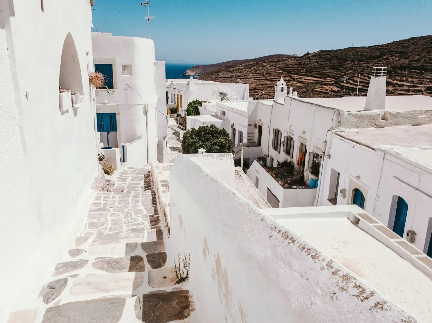 Whitewashed houses with blue doors along a stone-paved lane, terraced hills and sea beyond, Sifnos