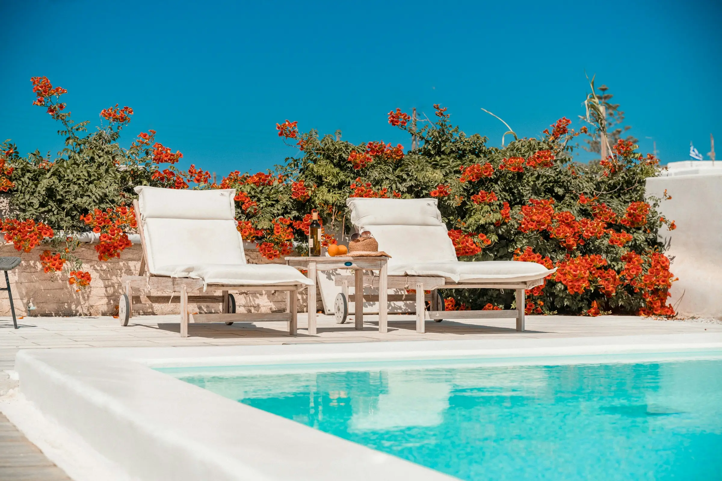 Cushioned sun loungers and a small table beside a turquoise pool, orange flowers behind, Mykonos