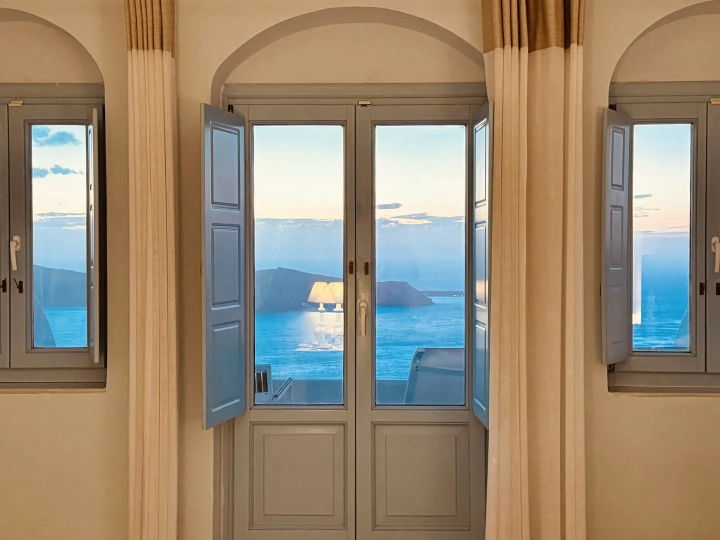 Hotel room in Santorini with arched windows, open blue shutters and linen curtains facing the sea at dusk
