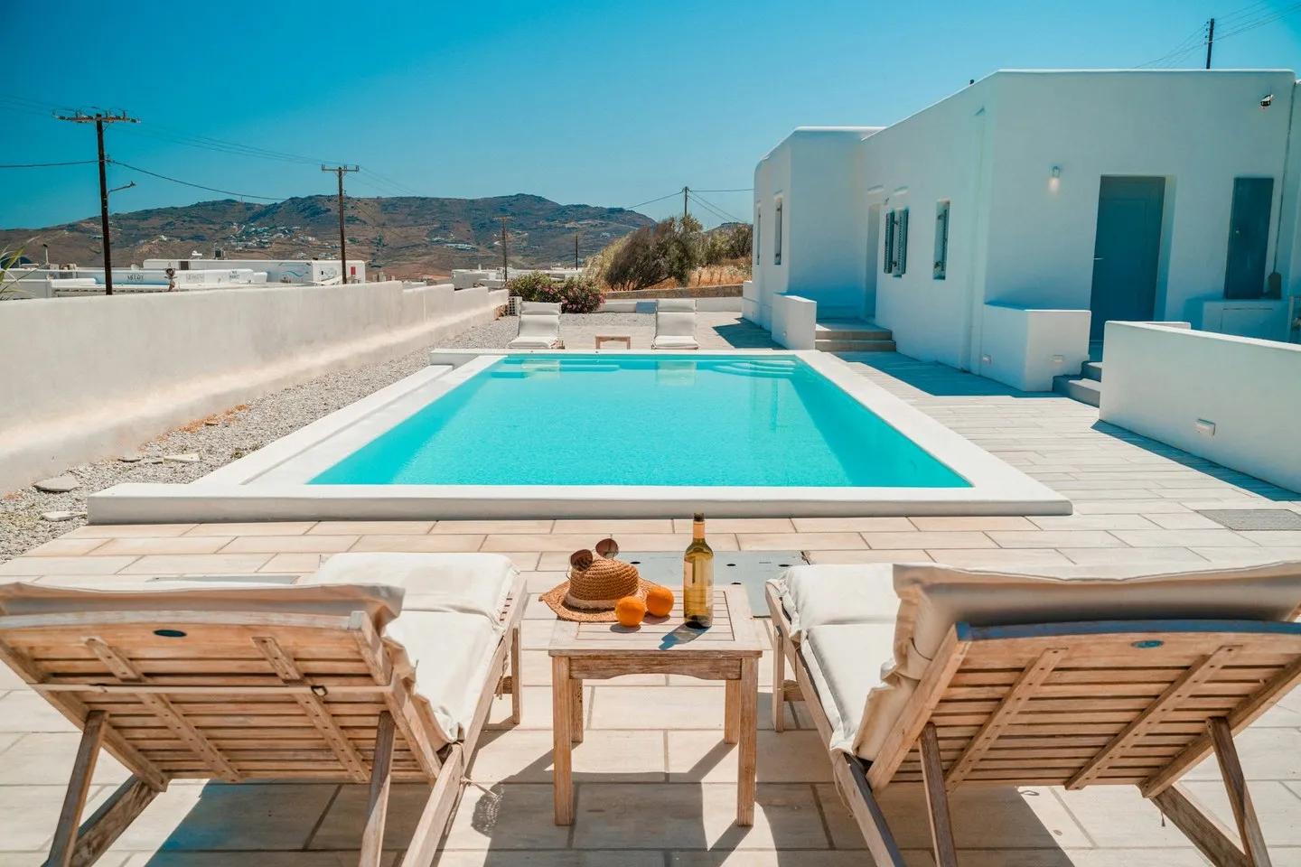 Mykonos pool deck with wooden sun loungers, a small table with a wine bottle and a whitewashed house