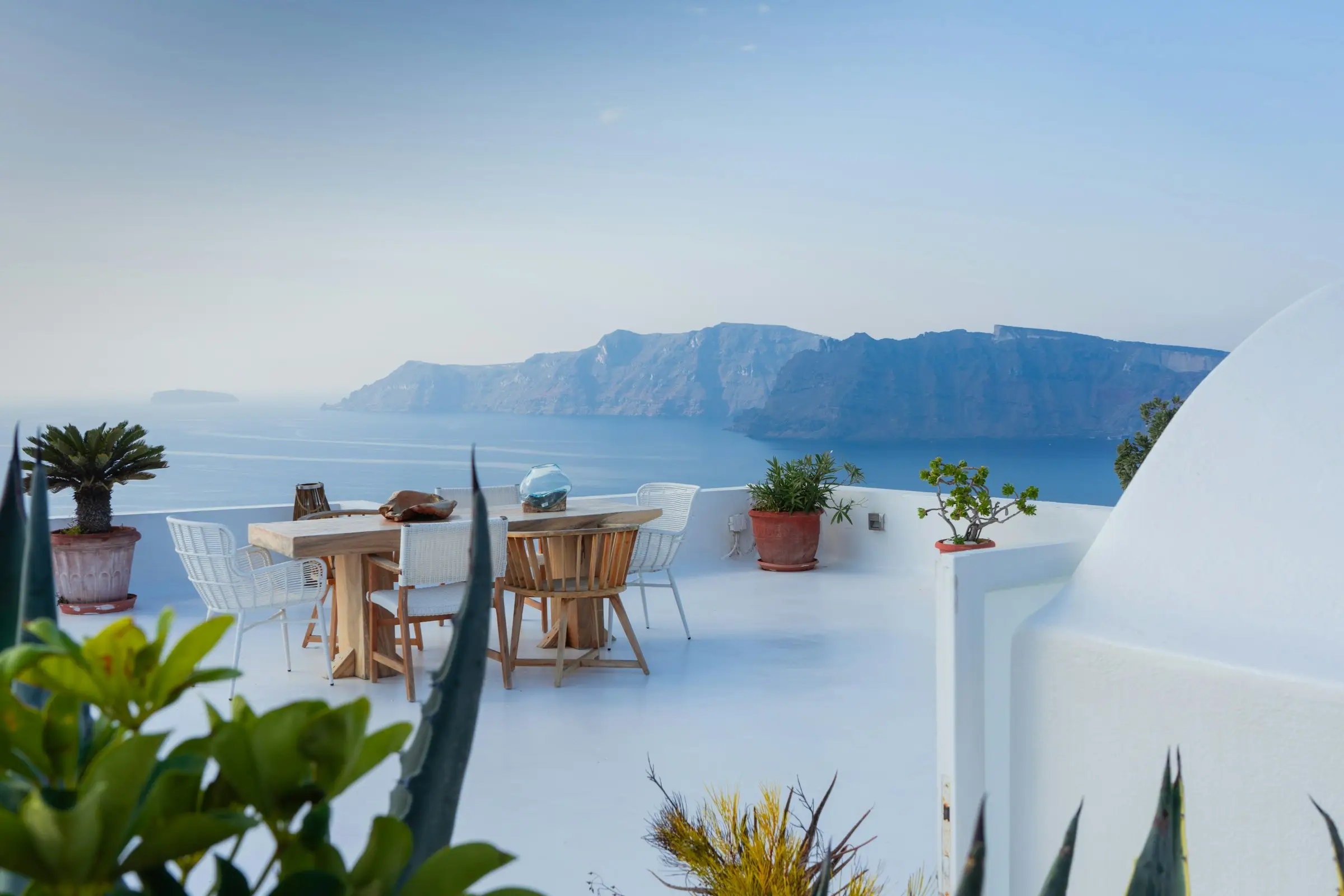 White terrace in Oia with a wooden table, chairs and potted plants overlooking the Santorini caldera