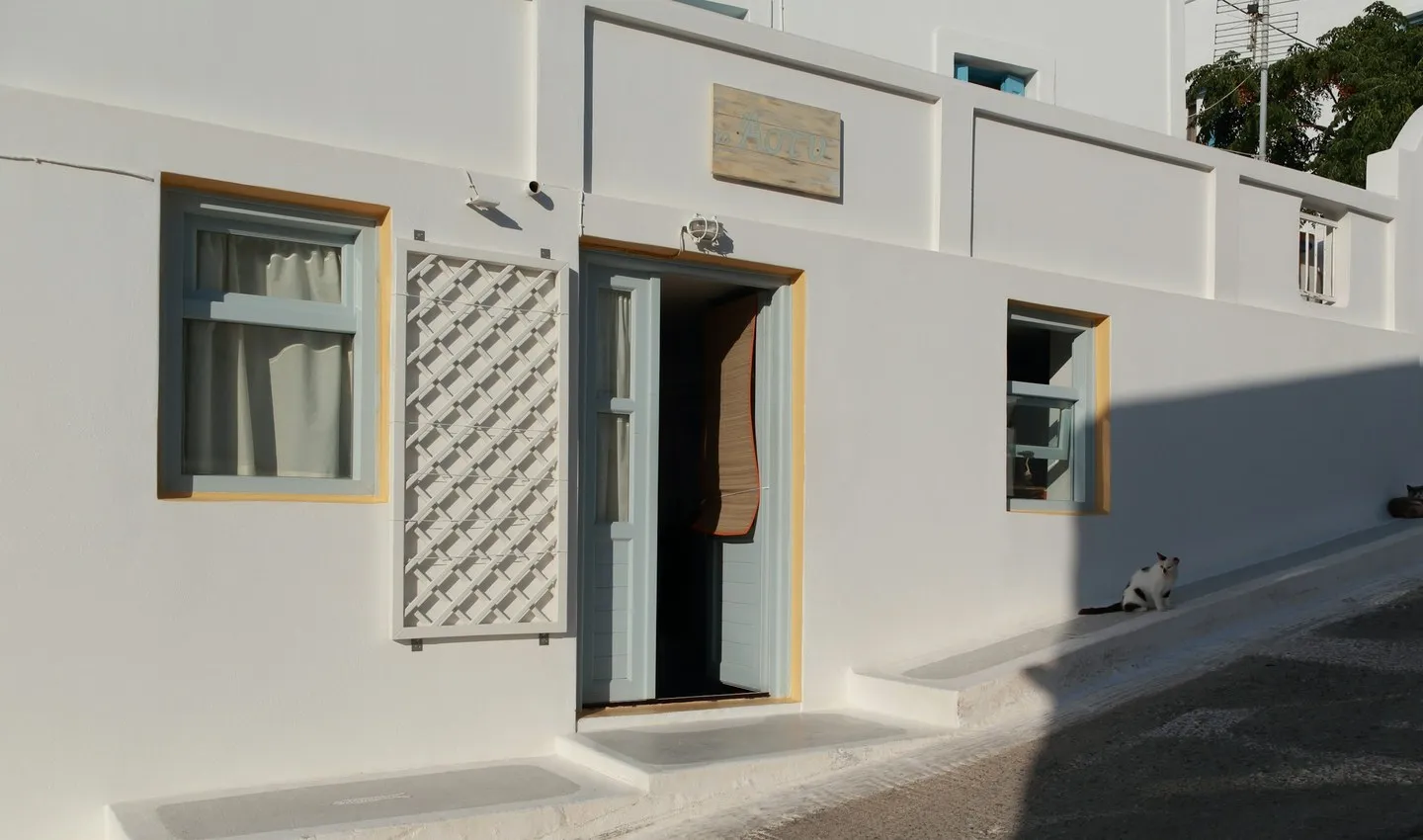 Whitewashed Astypalea building with an open blue door, two windows and a cat on the step