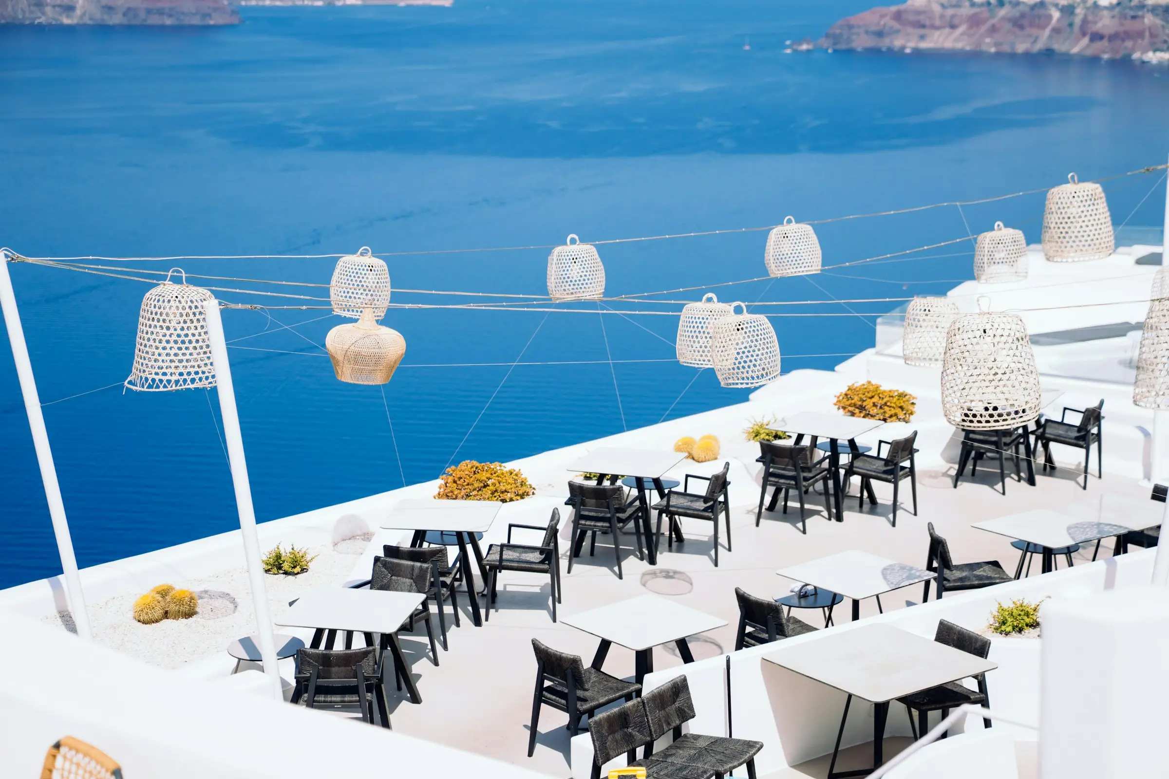 White restaurant terrace with dark chairs and woven lanterns above the blue sea in Santorini